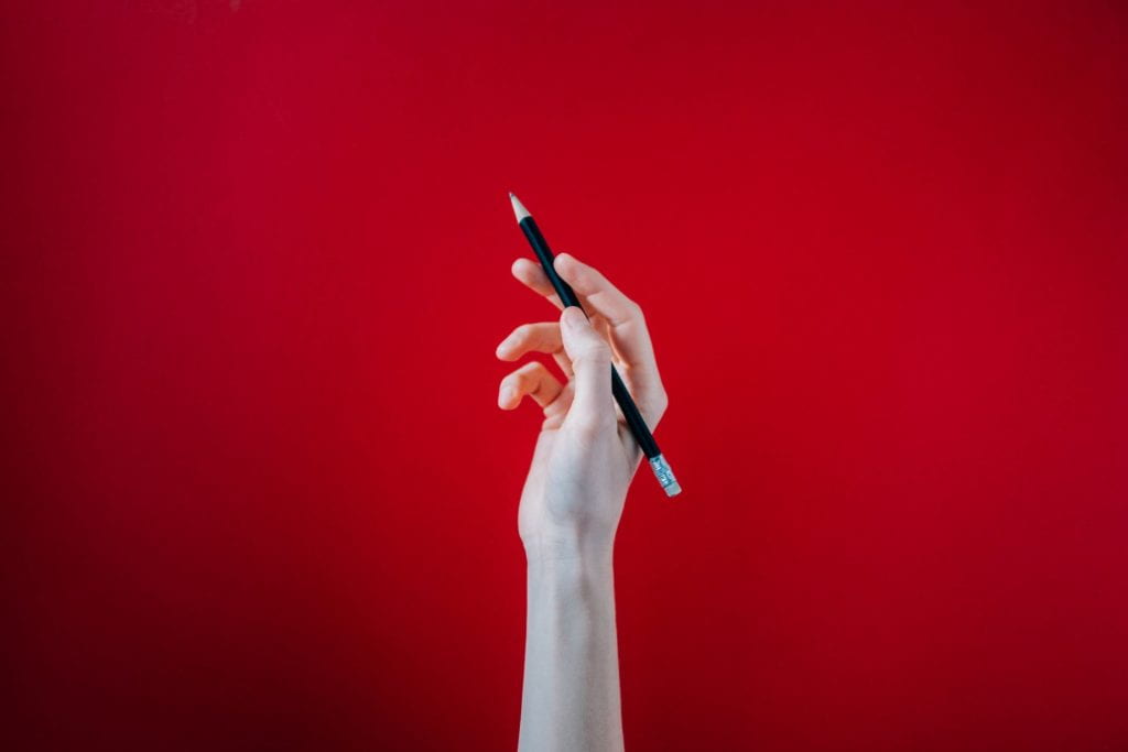 A white hand holds a pencil in front of a red background