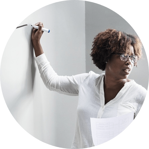 A Black woman in a white shirt is writing on a whiteboard while looking over her left shoulder.
